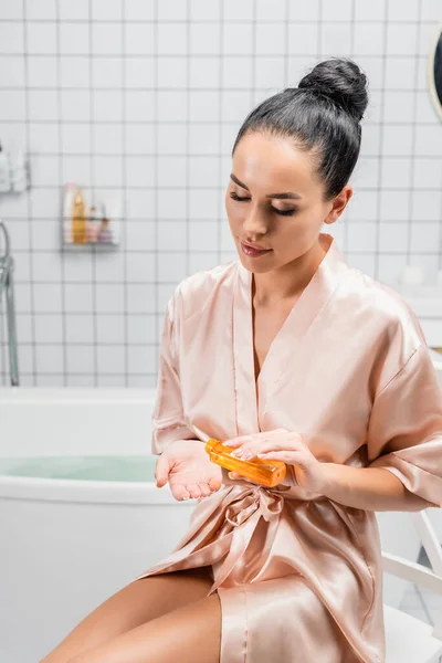 Молодая женщина в атласном халате заливает косметическое масло в ванную комнату — стоковое фото