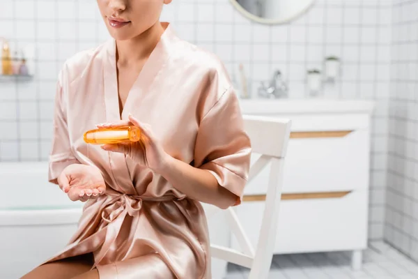 Обрезанный вид женщины в шелковом халате, наливающей косметическое масло в ванную комнату — стоковое фото
