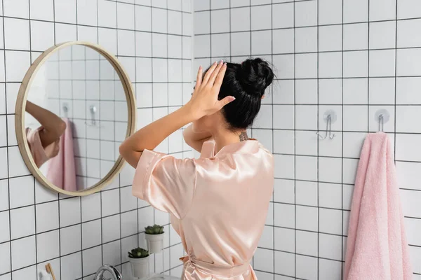 Jeune femme en satin peignoir toucher les cheveux près du miroir dans la salle de bain — Photo de stock