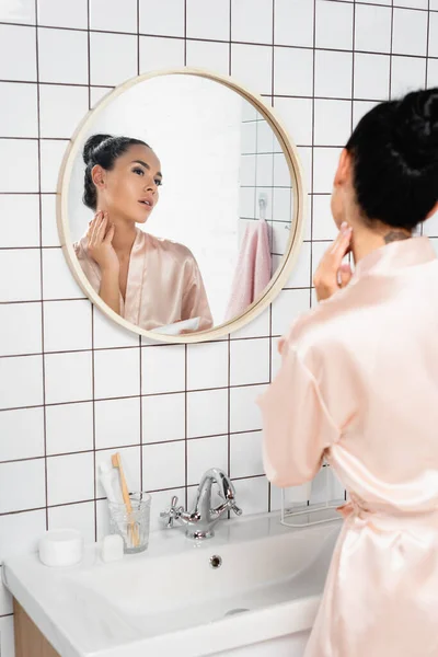 Mujer morena aplicando crema cosmética cerca del lavabo y espejo en el baño — Stock Photo