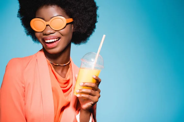 Mujer joven afroamericana feliz en traje elegante con jugo de naranja aislado sobre fondo azul - foto de stock