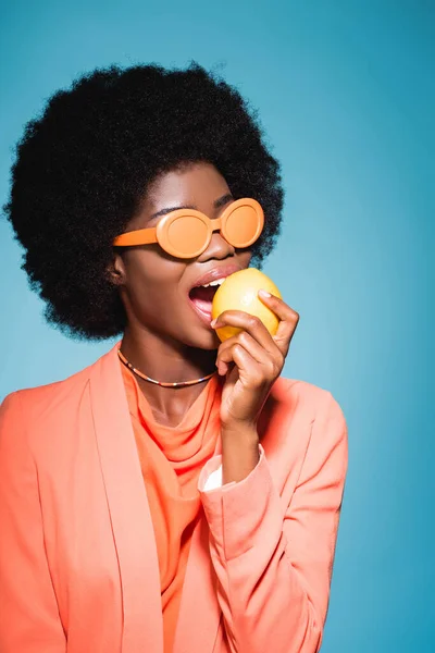Mujer joven afroamericana en naranja elegante traje mordiendo limón aislado sobre fondo azul - foto de stock