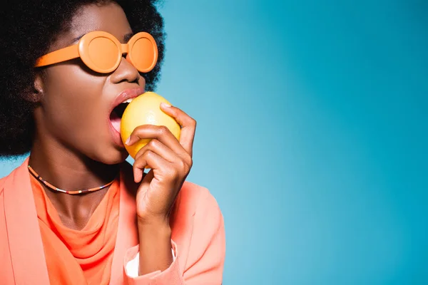 Афро-американська молода жінка в помаранчевому стильному вбранні кусає лимон ізольовано на синьому фоні — стокове фото