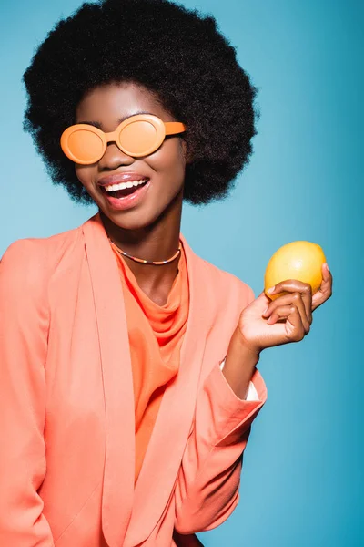 Mujer joven afroamericana feliz en traje elegante naranja con limón aislado sobre fondo azul - foto de stock