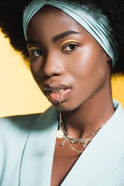 Afro-américaine jeune femme en tenue élégante bleue avec collier dans la bouche isolé sur jaune — Photo de stock