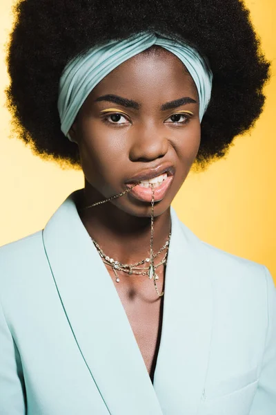 Afro-américaine jeune femme en tenue élégante bleue avec collier dans la bouche isolé sur jaune — Photo de stock