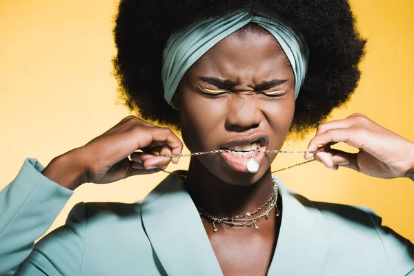 Африканская американская молодая женщина в синем стильном наряде с ожерельем во рту изолированы на желтый — стоковое фото