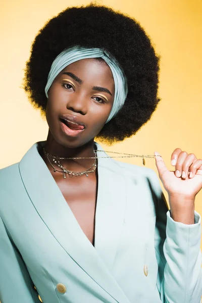 African american young woman in blue stylish outfit showing necklace isolated on yellow — Stock Photo