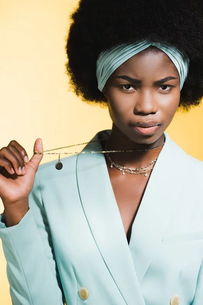 Seria africana americana joven mujer en azul elegante traje mostrando collar aislado en amarillo - foto de stock