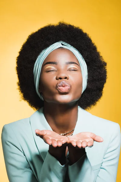 Afro americano jovem mulher no azul elegante roupa soprando beijo isolado no amarelo — Fotografia de Stock