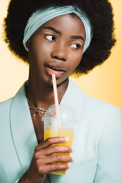 African american young woman in blue stylish outfit with orange juice isolated on yellow — Stock Photo