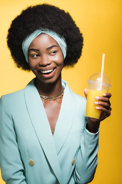 Jeune femme afro-américaine souriante dans une tenue élégante bleue avec du jus d'orange isolé sur jaune — Photo de stock