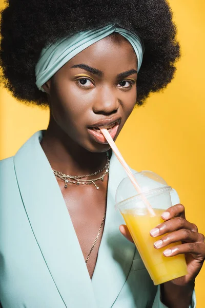 Brutal african american young woman in blue stylish outfit with orange juice isolated on yellow — Stock Photo