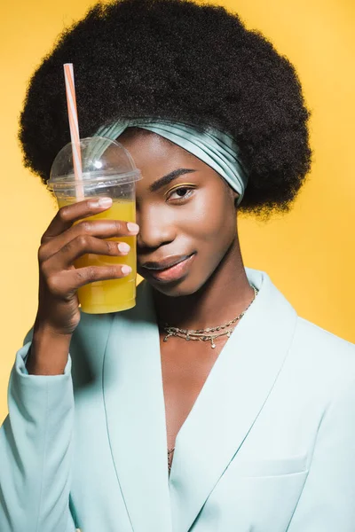 Afro-américaine jeune femme en tenue bleue stylée avec du jus d'orange isolé sur jaune — Photo de stock