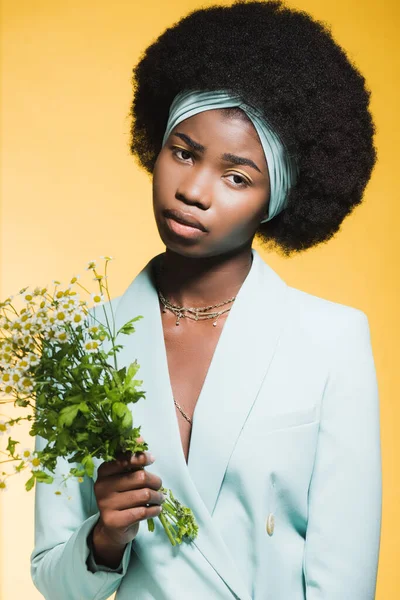 Афро-американська молода жінка в синьому стильному вбранні з букетом ромашки ізольовані на жовтому — стокове фото
