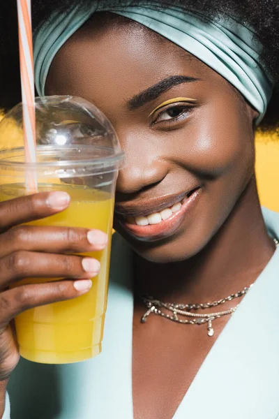 Close-up da jovem americana africana sorridente em roupa azul elegante com suco de laranja isolado no amarelo — Fotografia de Stock