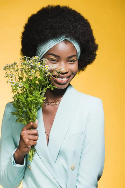 Sonriente mujer joven afroamericana en traje elegante azul con ramo de manzanilla aislado en amarillo - foto de stock