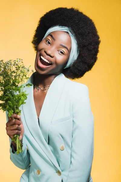 Happy african american young woman in blue stylish outfit with chamomile bouquet isolated on yellow — Stock Photo