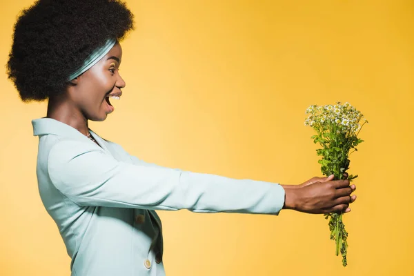 Vista laterale di felice donna africana americana in abito elegante blu con bouquet di camomilla isolato su giallo — Foto stock