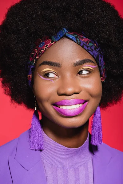 Smiling african american young woman in purple stylish outfit isolated on red — Stock Photo