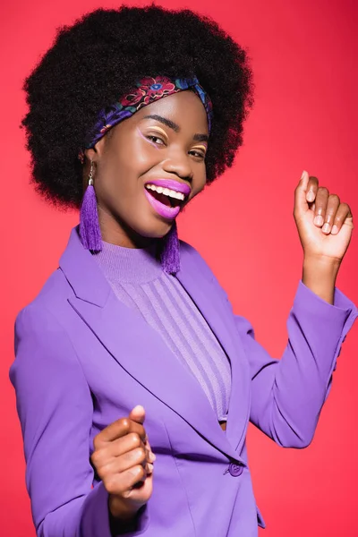 Счастливая африканская американская молодая женщина в фиолетовом стильном наряде, изолированном на красном — стоковое фото