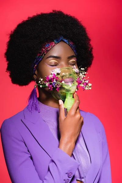 Afro-américaine jeune femme en violet tenue élégante avec des fleurs dans inhalateur isolé sur rouge — Photo de stock