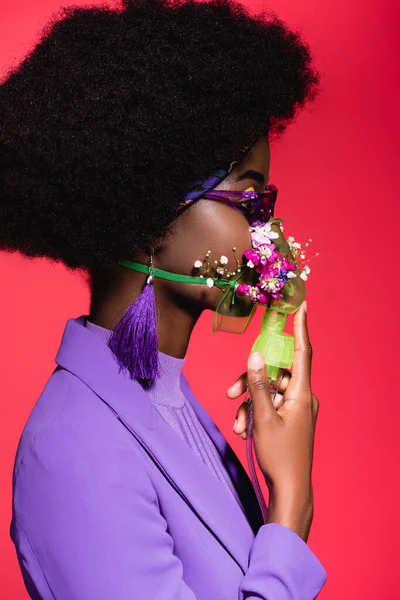 Vista laterale della giovane donna afroamericana in abito viola elegante con fiori in inalatore isolato su rosso — Foto stock