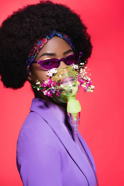 Афроамериканська молода жінка в фіолетовому стильному вбранні з квітами в інгаляторі ізольована на червоному — стокове фото