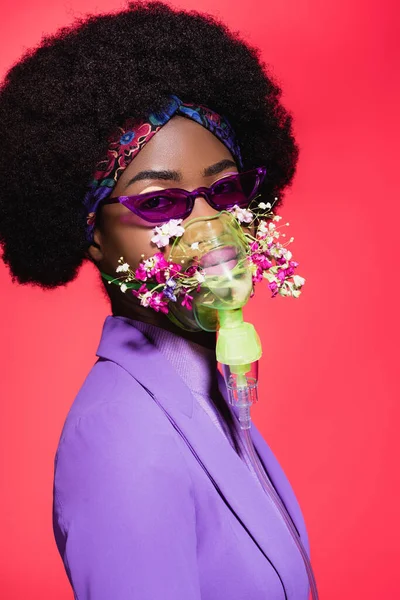 Mujer joven afroamericana en traje elegante púrpura con flores en inhalador aislado en rojo - foto de stock