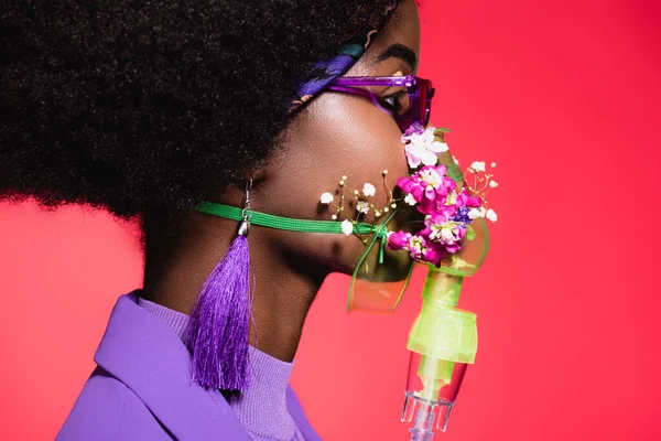 Vista laterale della giovane donna afroamericana in abito viola elegante con fiori in inalatore isolato su rosso — Foto stock