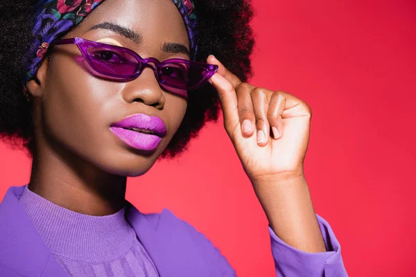 Afro-américaine jeune femme en violet tenue élégante et lunettes de soleil isolées sur rouge — Photo de stock