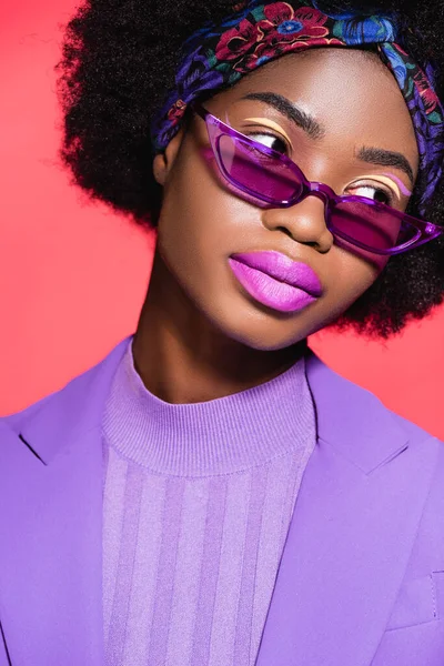 Afro-américaine jeune femme en violet tenue élégante et lunettes de soleil isolées sur rouge — Stock Photo