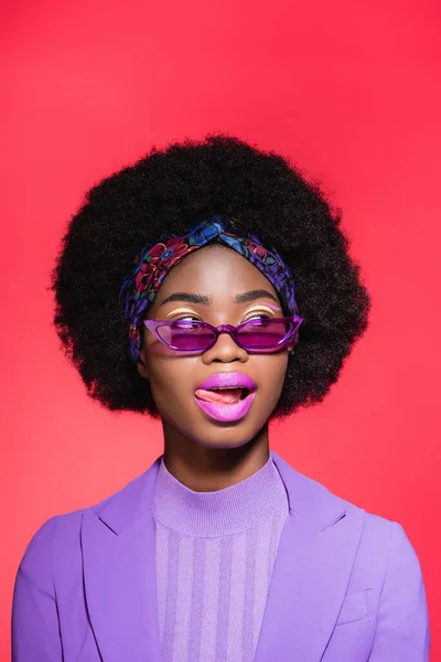 Afro-américaine jeune femme en violet tenue élégante et lunettes de soleil montrant la langue isolée sur rouge — Photo de stock