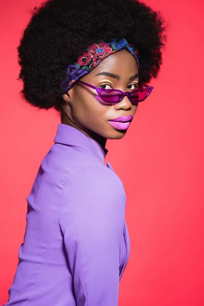 Вид збоку афроамериканської молодої жінки в фіолетовому стильному вбранні та сонцезахисних окулярах ізольовані на червоному — стокове фото
