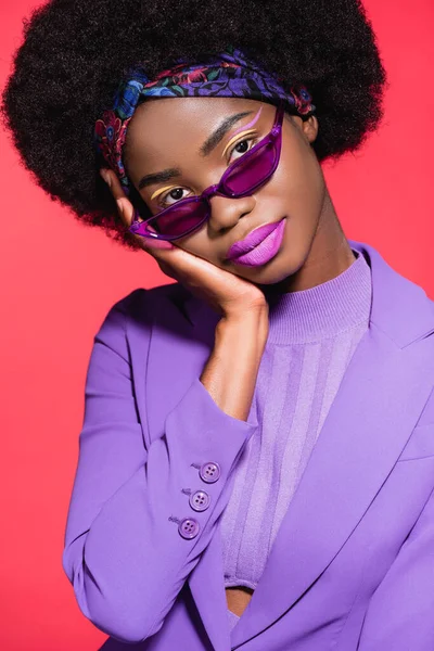 Mujer joven afroamericana en traje elegante púrpura y gafas de sol aisladas en rojo - foto de stock
