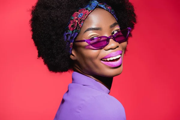 Happy african american young woman in purple stylish outfit and sunglasses isolated on red — Stock Photo