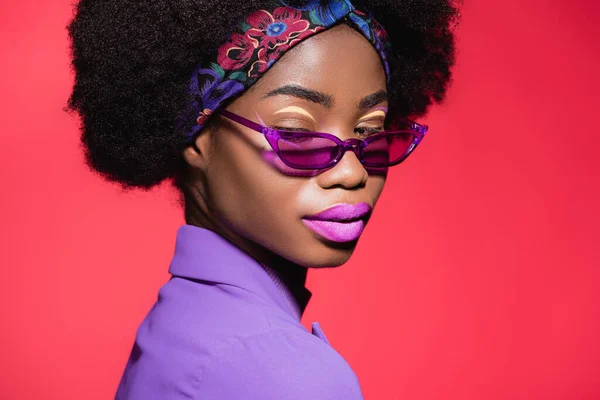 Afro-américaine jeune femme en violet tenue élégante et lunettes de soleil isolées sur rouge — Photo de stock