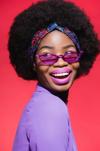 Heureux afro-américaine jeune femme en violet tenue élégante et lunettes de soleil isolées sur rouge — Photo de stock