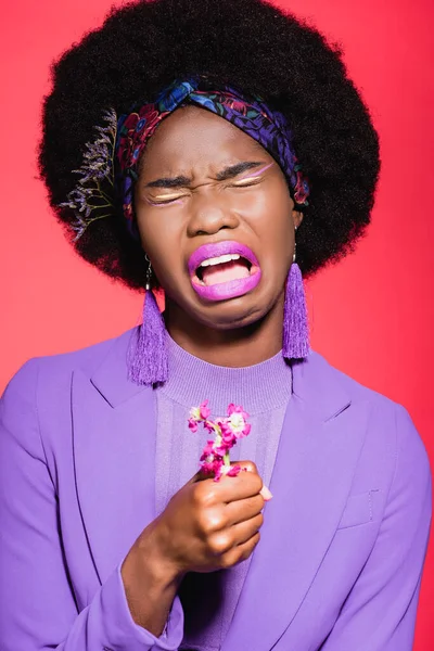 Triste afro-americana jovem mulher em roupa elegante roxo com flor chorando isolado no vermelho — Fotografia de Stock