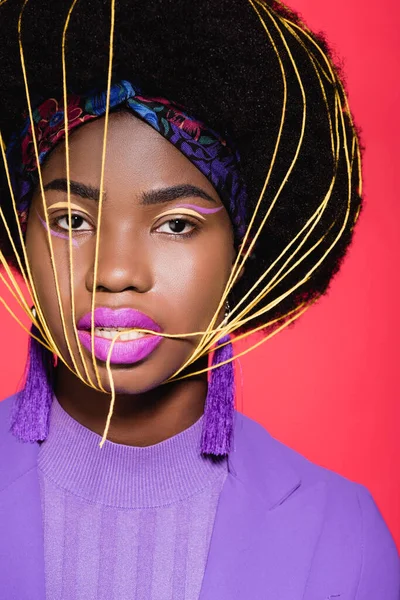 Mujer joven afroamericana en traje elegante púrpura con cuerdas amarillas en la cara aislada en rojo - foto de stock