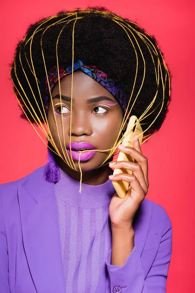 African american young woman in purple stylish outfit with yellow strings on face holding smartphone isolated on red — Stock Photo
