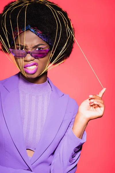 African american young woman in purple stylish outfit and sunglasses with yellow strings on face isolated on red — Stock Photo