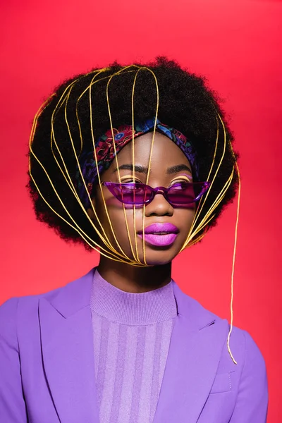 Afro-américaine jeune femme en costume violet élégant et des lunettes de soleil avec des cordes jaunes sur le visage isolé sur le rouge — Photo de stock