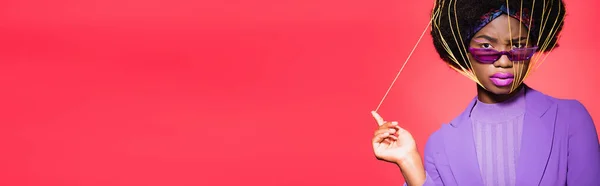 Giovane donna afroamericana in abito viola elegante e occhiali da sole con stringhe gialle sul viso isolato su rosso, banner — Foto stock