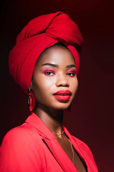 Mujer joven afroamericana en traje elegante rojo y turbante aislado en negro - foto de stock