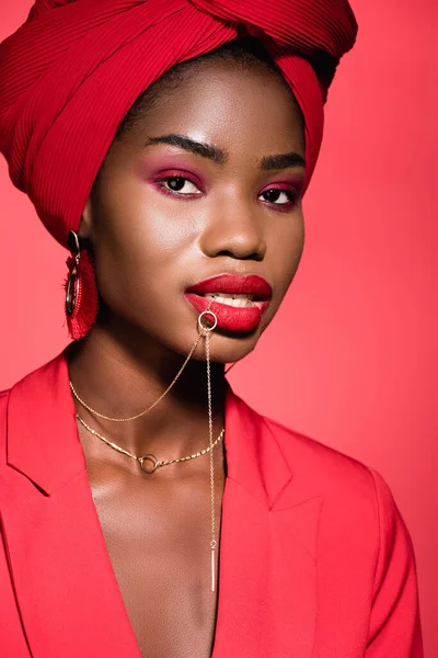 Africano americano jovem mulher na roupa elegante e turbante segurando colar na boca isolado no vermelho — Fotografia de Stock