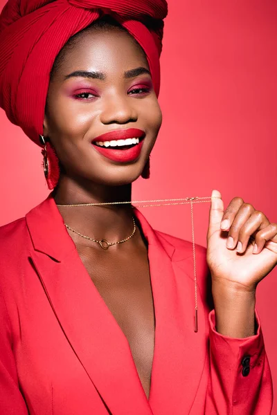 Souriant afro-américaine jeune femme en tenue élégante et turban tenant collier isolé sur rouge — Photo de stock