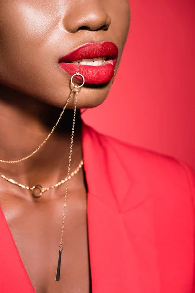 Cropped view of african american young woman in stylish outfit holding necklace in mouth isolated on red — Stock Photo