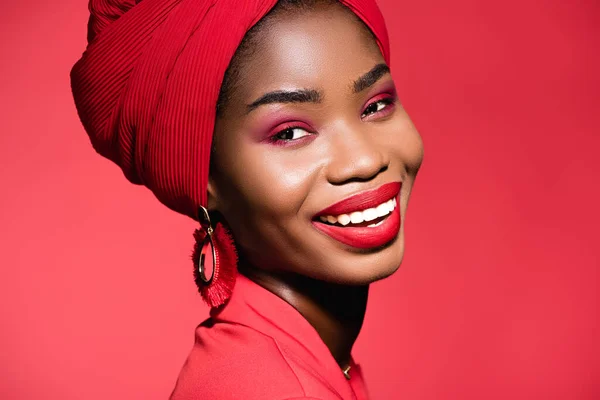 Улыбающаяся африканская американская молодая женщина в стильном наряде и тюрбане, изолированном на красном — стоковое фото