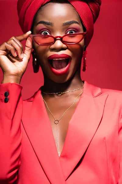 Excited african american young woman in stylish outfit, sunglasses and turban isolated on red — Stock Photo